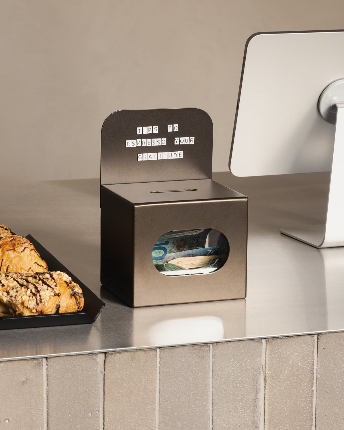 Bronze FOLD Tip Box by Made of Tomorrow on a café counter beside pastries and a payment terminal
